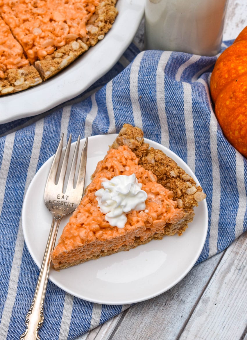 a slice of a pumpkin pie rice krispie treat topped with whipped cream on a small white dessert plate with a silver fork resting on the side