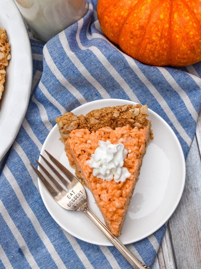 a slice of a pumpkin pie rice krispie treat topped with whipped cream on a small white dessert plate with a silver fork resting on the side