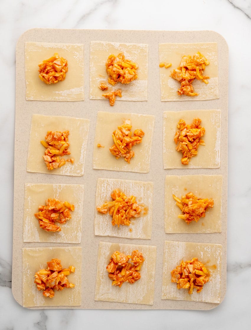 spoon fulls of creamy buffalo chicken filling on wonton wrappers arranged in rows on a cutting board