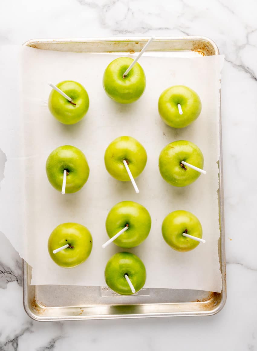 lollipop sticks stuck in granny smith apples on a parchment lined baking sheet pan