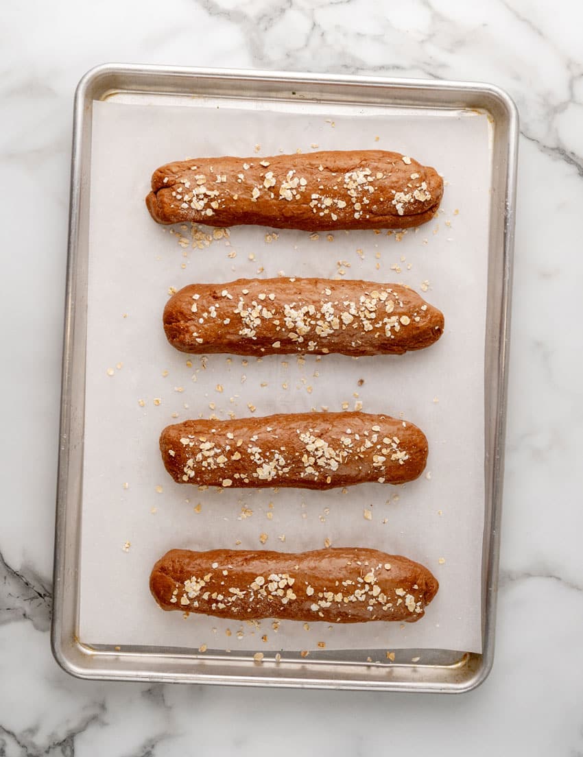 homemade logs of restaurant style brown bread topped with rolled oats arranged in a row on a parchment paper lined baking tray