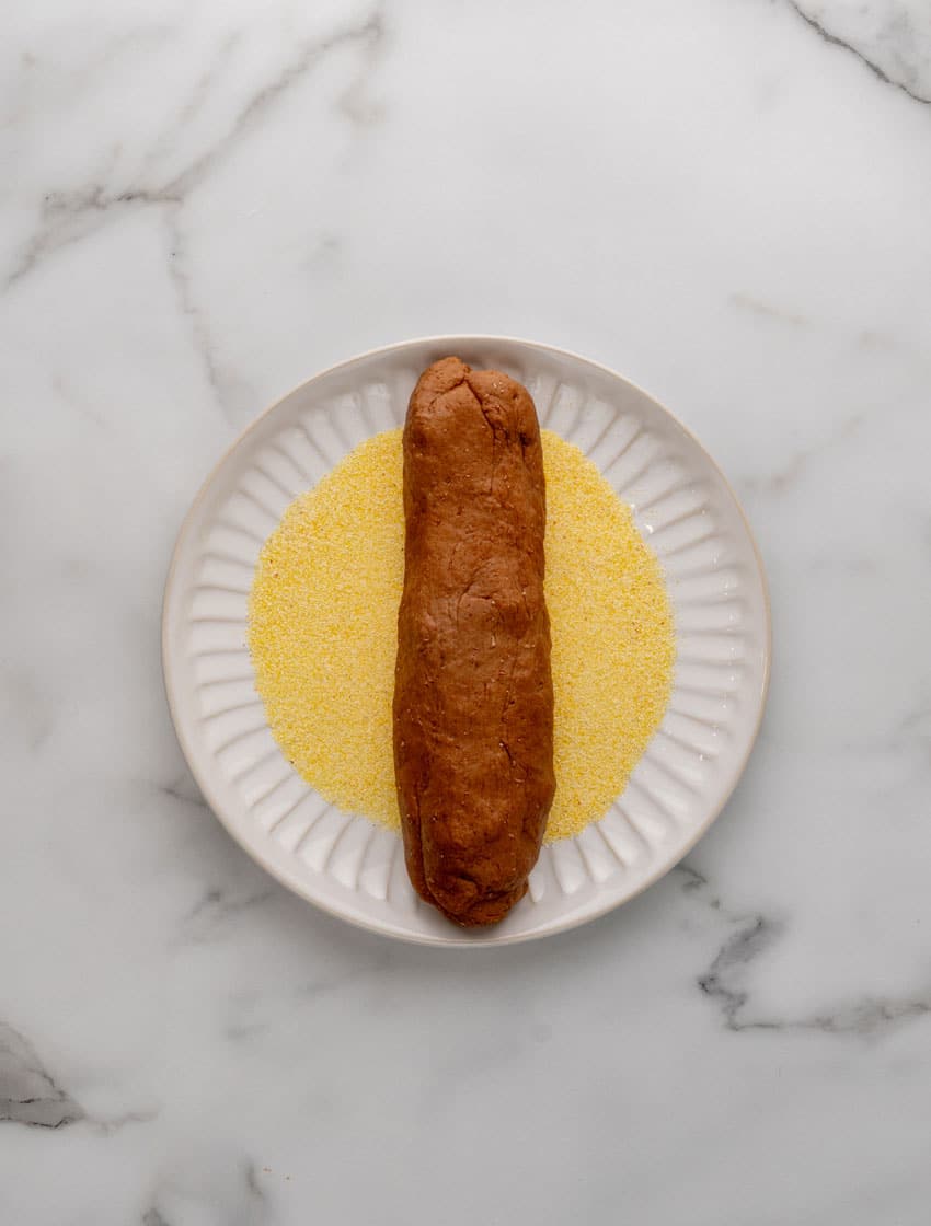 a roll of copycat cheesecake factory brown bread resting on a plate filled with cornmeal