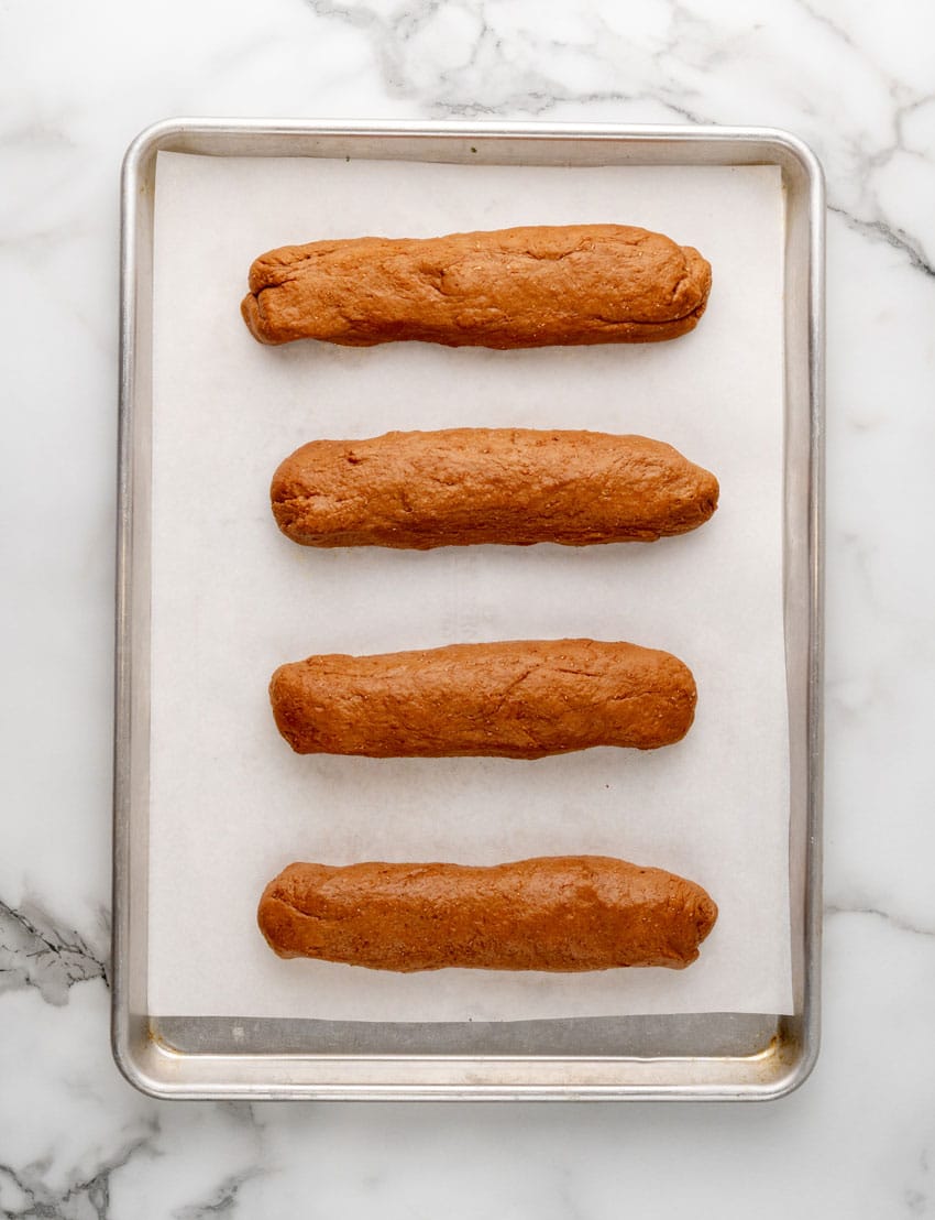 homemade logs of restaurant style brown bread coated in cornmeal arranged in a row on a parchment paper lined baking tray
