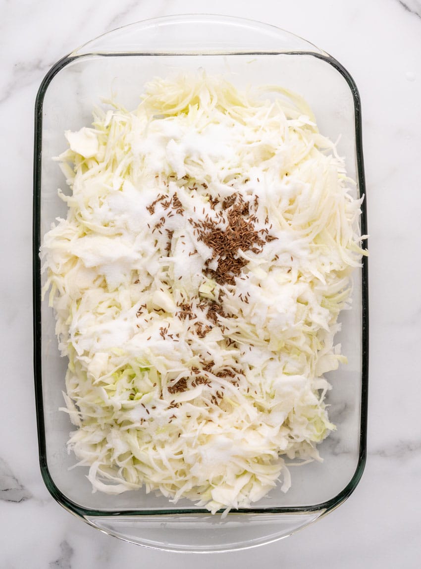 salt and caraway seeds spread out overtop thinly sliced fresh cabbage in a large glass baking dish