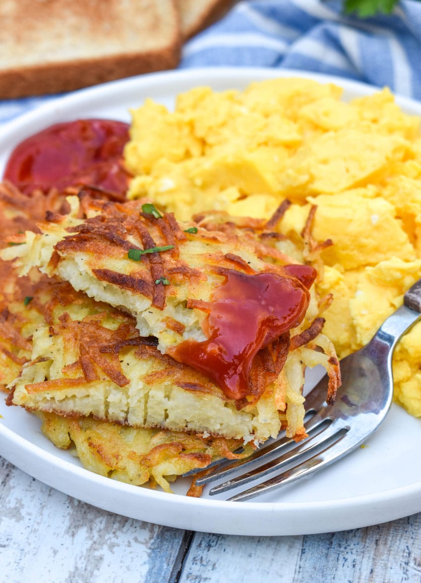crispy homemade hash brown patties topped with ketchup next to scrambled eggs on a white plate