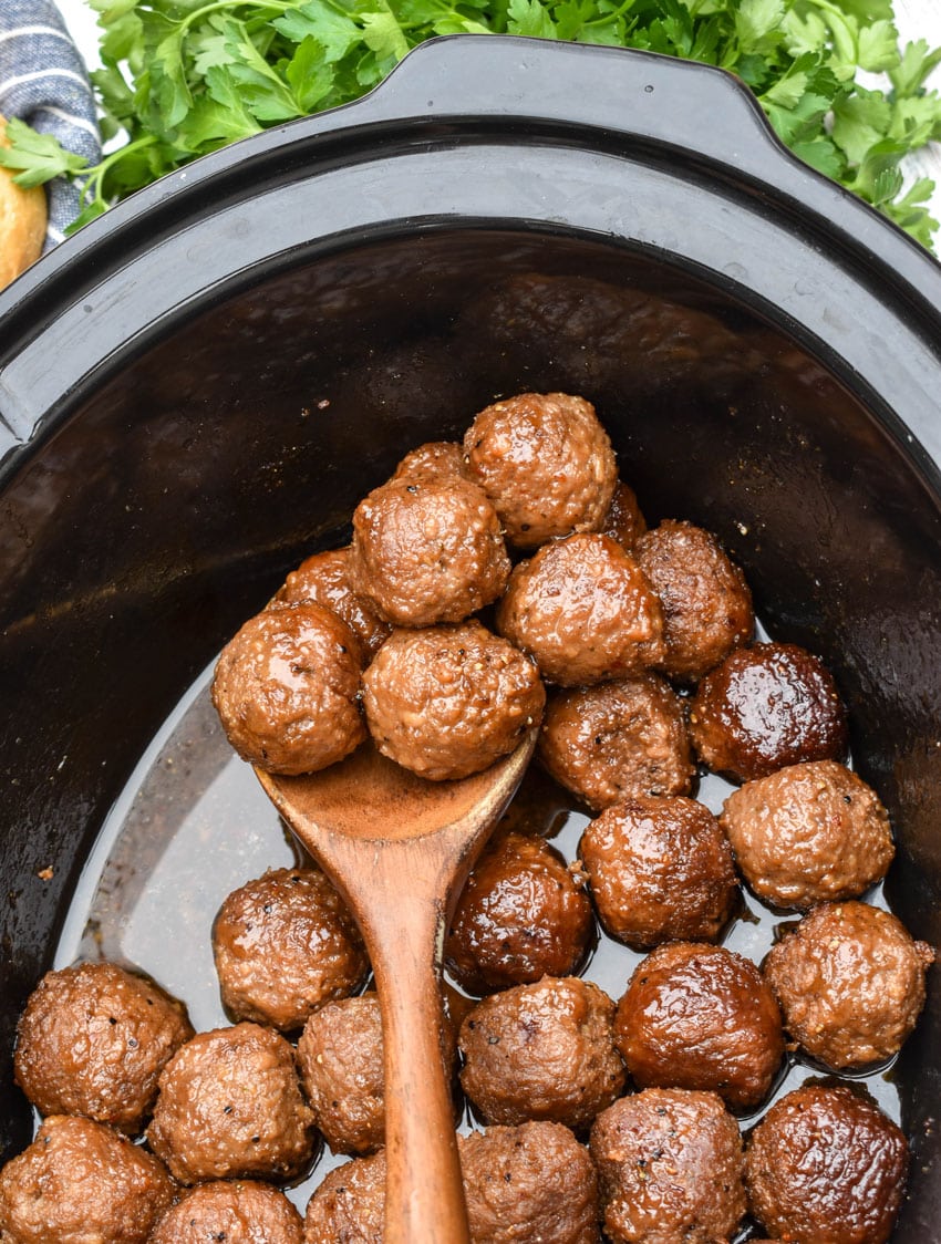 a wooden spoon scooping brown sugar meatballs out of the black bowl of a slow cooker