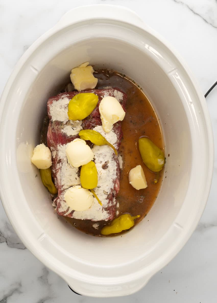 a raw chuck roast topped with ranch dressing mix, au jus gravy mix, pats of butter, and pepperoncini peppers in the bowl of a white slow cooker