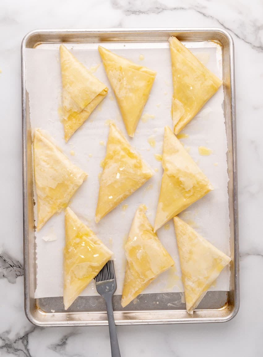 butter brushed pieces of homemade spanakopita arranged in rows on a parchment paper lined baking sheet